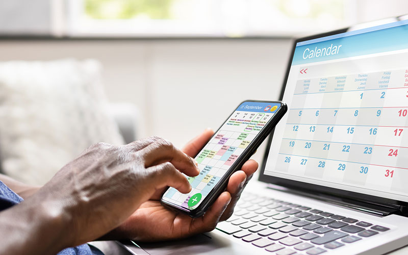 close up of a hand holding a phone with a calendar on screen with a laptop calendar in the background