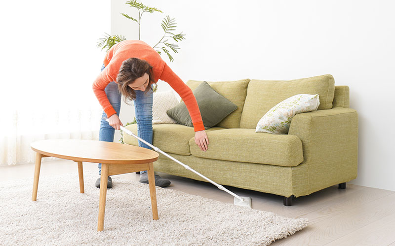 a woman reaching to mop underneath a sofa in a bright, well lit living room